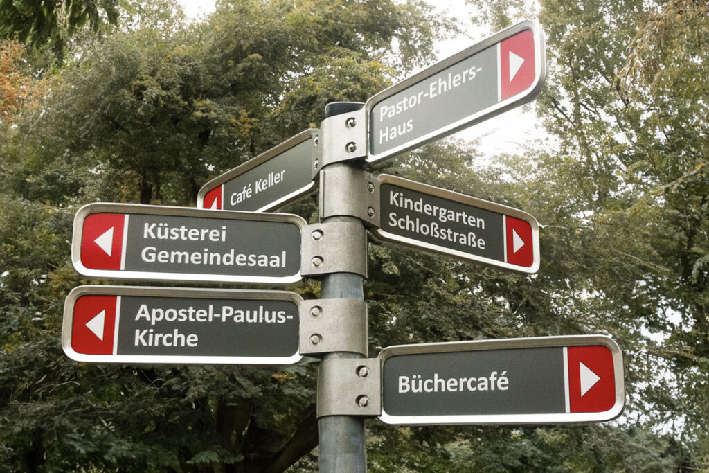 Mehrfachwegweiser-Stele einer Berliner Pfarrei, graue, metallgerahmte Hinweisschilder mit weißen Pfeilen auf rotem Hintergrund an der Spitze.